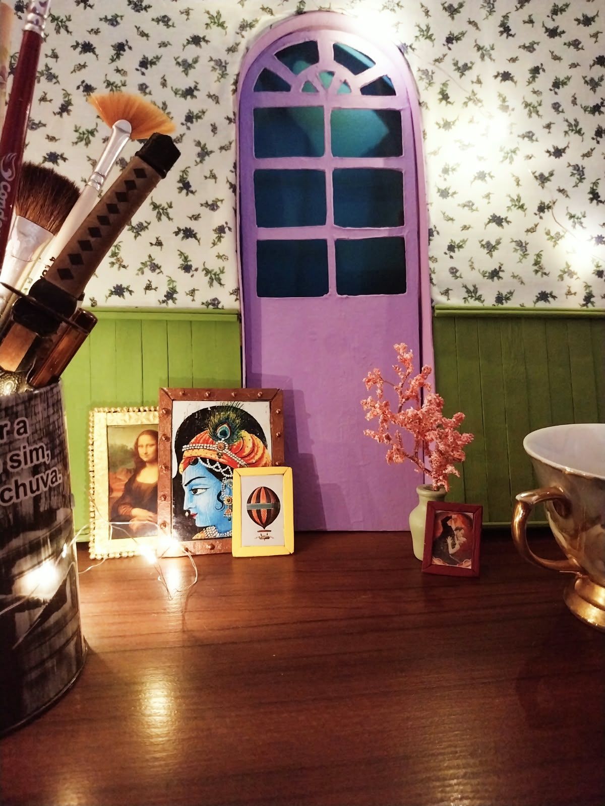Bancada artística do atelier Madame Blum com pincéis, iluminação suave, portal lilás e obras em miniatura